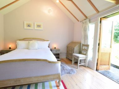 A bedroom with a wooden bed white bedding two bedside tables lamps a chair a desk a rug and an open door to outside at The White House with Studio in Wortwell