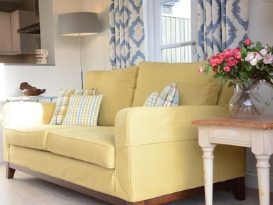 A yellow sofa with patterned cushions next to a small table with a vase of flowers and a floor lamp behind at Mellows Thorpeness in Thorpeness