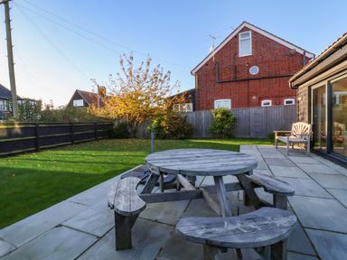 A backyard with a round wooden picnic table on a stone patio next to a wooden bench and a lawn at Mellows Thorpeness in Thorpeness