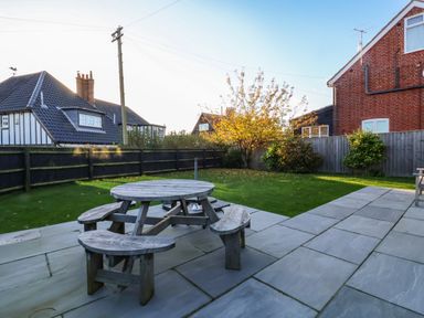 A backyard with a round wooden picnic table on a tiled patio and grass with trees and bushes at Mellows Thorpeness in Thorpeness