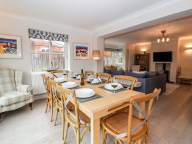 A dining area with a wooden table set with plates and wine glasses next to a living room with blue sofas and a wall-mounted television at Mellows Thorpeness in Thorpeness
