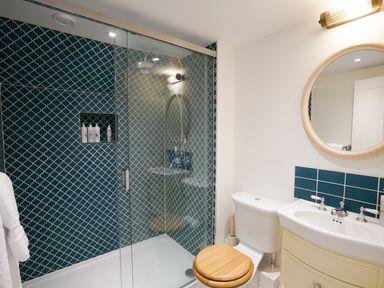 A bathroom with a glass shower enclosure blue tiled wall a white toilet with wooden seat and a sink with a round mirror and blue tile backsplash at Shingle Skies Aldeburgh