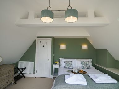 A bedroom with a double bed with pillows and a tray, a green accent wall with lights, a chest of drawers, and a door at Shingle Skies Aldeburgh