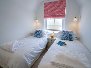 A bedroom with two single beds white bedding blue patterned pillows a bedside table with drinks and croissants and a red patterned window blind at Shingle Skies Aldeburgh