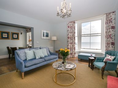 A living room with a blue sofa, armchair, marble coffee table with flowers, bay window, and sea view at Shingle Skies Aldeburgh