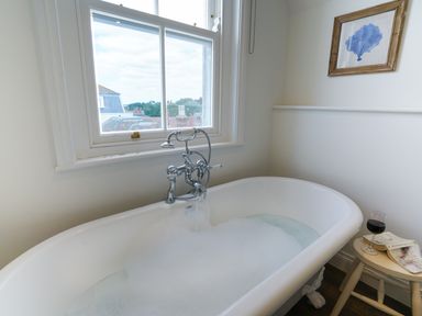 A bathroom with a clawfoot bathtub filled with bubble bath water under a window and a small side table with a glass of wine and books at Shingle Skies Aldeburgh