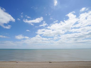 A beach with two people sitting near the water and a partly cloudy sky at Shingle Skies Aldeburgh