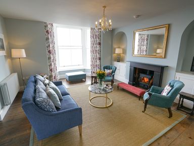 A living room with a blue sofa, two teal armchairs, a round coffee table, a fireplace with a gold mirror above it, and a large window at Shingle Skies, Aldeburgh