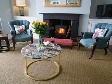 A living room with a stone coffee table yellow flowers in a vase a burning fireplace and two blue armchairs at Shingle Skies Aldeburgh