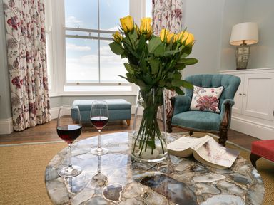 A living room with yellow roses in a glass vase red wine glasses and an open book on a stone coffee table at Shingle Skies Aldeburgh
