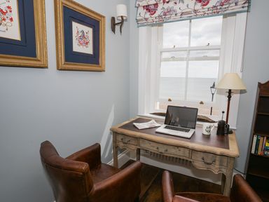 A study corner with a wooden desk, two leather chairs, a laptop, a newspaper, a lamp, and a window overlooking the sea at Shingle Skies, Aldeburgh