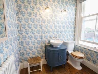 A bathroom with a blue cabinet and sink a toilet with a wooden seat a small stool and a window at Shingle Skies Aldeburgh