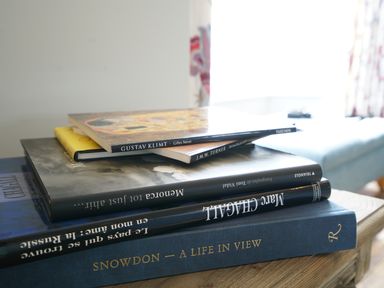 A stack of books on a wooden table with a couch and window in the background at Shingle Skies Aldeburgh