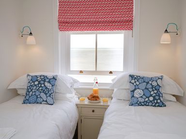 A bedroom with two single beds with white bedding and blue patterned pillows a window with a red and white blind and a nightstand with juice and croissants at Shingle Skies Aldeburgh