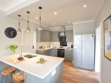 A kitchen with grey cabinets white countertops a silver refrigerator wooden stools and pendant lights at The Round House in Easton near Framlingham