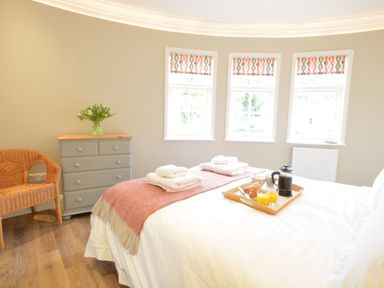 A bedroom with a bed a tray with breakfast a chest of drawers wicker chair and three windows at The Round House in Easton near Framlingham