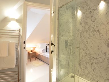 A bathroom with a glass shower and towels on a rack viewed towards a bedroom with a bed and bedside table at The Round House in Easton near Framlingham