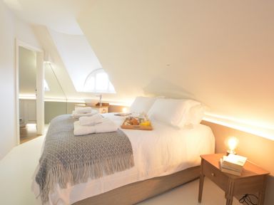 A bedroom with a bed with white bedding and folded towels on it a wooden nightstand with a lamp and books and a small window in the background at The Round House in Easton near Framlingham