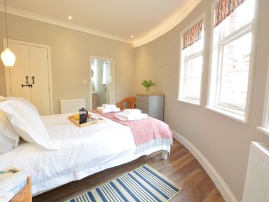 A bedroom with a bed, bedside table with books, a striped rug, dresser with flowers, and windows at The Round House in Easton near Framlingham