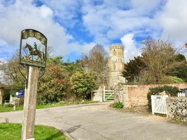 A village road with a wooden signpost showing a horse rider and dogs with a round clock tower and trees in the background at Easton near Framlingham