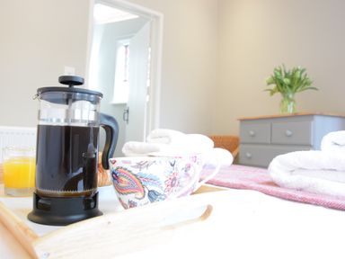 A tray with a French press coffee pot two cups and a glass of orange juice on a bed with towels and a gray dresser with flowers at The Round House in Easton near Framlingham