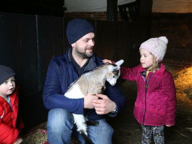 A man holding a goat with two children petting it inside a barn with hay on the floor at The Round House in Easton near Framlingham