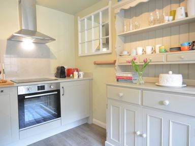 A kitchen with an oven and stove next to a cupboard with shelves holding cups and glasses with flowers on the countertop at Church Farm Cottage Dunwich Near Westleton