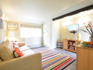 A living room with a beige sofa and orange pillows a striped rug a wooden cabinet with books and a yellow vase a wall-mounted television and a window at Church Farm Cottage Dunwich Near Westleton