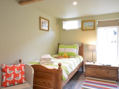 A bedroom with a single wooden bed, a striped chair with a bird cushion, a wooden chest, and framed artworks on the walls at Church Farm Cottage Dunwich Near Westleton
