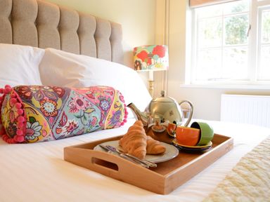 A breakfast tray with a teapot, croissants, and colorful cups on a bed with a patterned bolster pillow and a window in the background at Church Farm Cottage Dunwich Near Westleton