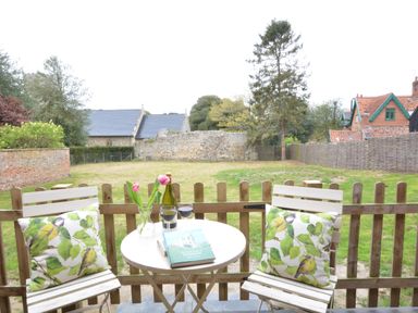 A small outdoor patio with two wooden chairs with bird print cushions a round table with a book wine glasses and tulips overlooking a fenced grassy yard and stone buildings at Church Farm Cottage Dunwich Near Westleton