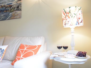 A corner of a living room with a white sofa with two pillows and a white side table with a lamp two glasses of red wine a magazine and a plate of grapes at Church Farm Cottage Dunwich Near Westleton