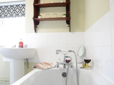 A bathroom with a white sink and bathtub with a glass of red wine and a small mirror on the tub edge at Church Farm Cottage Dunwich Near Westleton