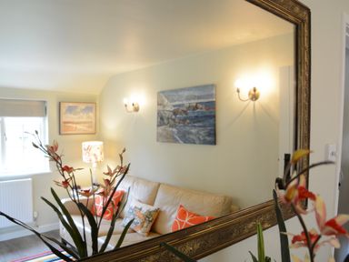 A reflection in a mirror showing a living room with a beige sofa with orange and patterned cushions a lamp a plant and paintings on the wall at Church Farm Cottage Dunwich Near Westleton