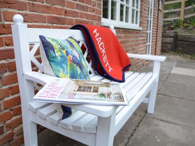 A white wooden bench with a colorful parrot pillow a red Hackett blanket and a magazine outside against a brick wall at Church Farm Cottage Dunwich Near Westleton