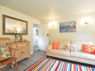 A living room with a cream sofa with orange and patterned cushions a striped rug a wooden chair with a pineapple cushion a wooden cabinet with fruit and flowers and a large mirror on the wall at Church Farm Cottage Dunwich Near Westleton