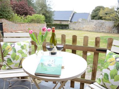 A small round outdoor table with a book and two glasses of red wine between two wooden chairs with floral cushions at Church Farm Cottage Dunwich Near Westleton