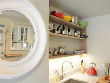 A kitchen corner with wooden shelves holding bowls pots plates and kettles near a sink with a red kettle at Church Farm Cottage Dunwich Near Westleton