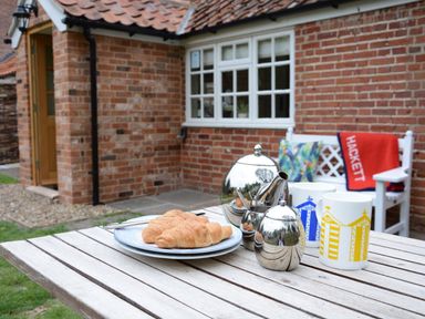 A wooden outdoor table with croissants on plates a teapot sugar pot two mugs a white bench with cushions and a red blanket on a patio next to a brick house at Church Farm Cottage Dunwich Near Westleton