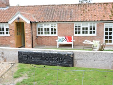 A brick cottage with a tiled roof and open wooden door behind a gate with a sign that says The Cottage at Church Farm Cottage Dunwich Near Westleton