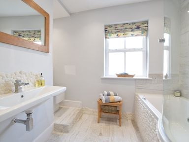 A bathroom with a sink mirror a small wooden stool with folded towels a window and a bathtub at 53 Tern Cottage Aldeburgh