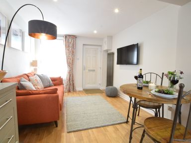 A living room with an orange sofa, a round wooden dining table with two chairs, a wall-mounted tv, and a floor lamp at 53 Tern Cottage, Aldeburgh