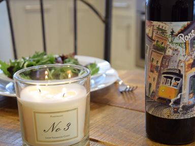 A lit candle next to a plate of salad and a bottle with a colorful label on a wooden table at 53 Tern Cottage in Aldeburgh