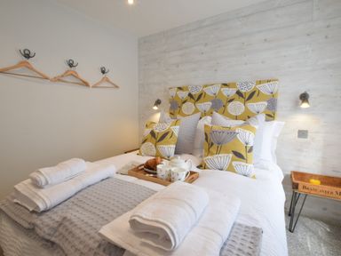 A bedroom with a bed with patterned cushions towels on a gray blanket a wooden tray with tea and croissants and wooden hangers on the wall at 53 Tern Cottage Aldeburgh