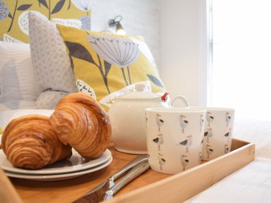 A breakfast tray with croissants teapot and cups on a bed with patterned pillows at 53 Tern Cottage Aldeburgh