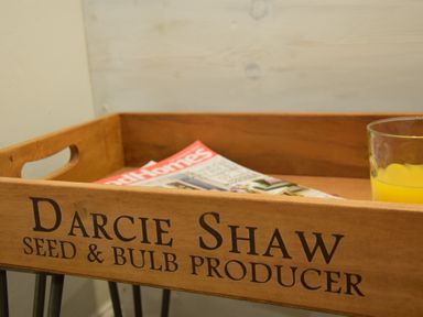 A wooden tray with the words Darcie Shaw Seed and Bulb Producer containing a magazine and a glass of orange juice