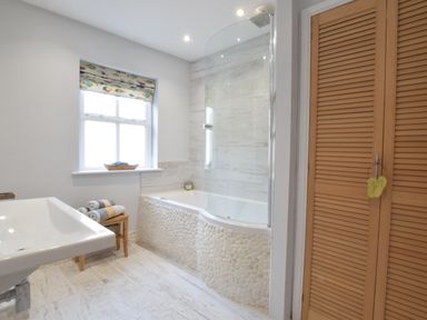 A bathroom with a bathtub, sink, wooden bench with towels, window with patterned blind, and wooden louvered doors at 53 Tern Cottage Aldeburgh