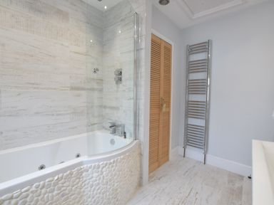 A bathroom with a bathtub surrounded by textured tiles a glass shower screen wooden louvered doors and a metal towel radiator at 53 Tern Cottage Aldeburgh