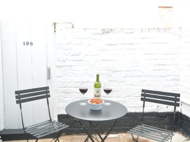A small outdoor seating area with two black metal chairs and a round black table with wine glasses a bottle and a bowl of strawberries at 51 Tern Cottage Aldeburgh