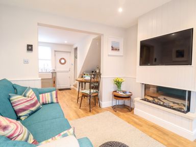 A living room with a teal sofa and colorful pillows a wall mounted TV and fireplace a small dining table and kitchen visible at 51 Tern Cottage Aldeburgh